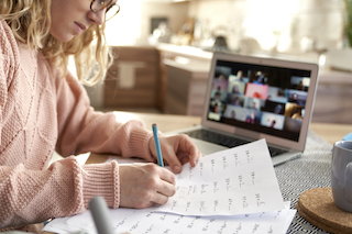 Student writing notes while sitting in front of a computer with a smartphone next to her